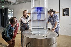 Drei Erwachsene beobachten einen Zylinder mit Wasser im 'Blauen Wunder' im Hexenwasser Söll.