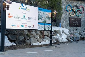 Informationsschild zum Mountain Travel Symposium mit Partnerlogos vor einer Steinwand mit olympischen Ringen und der Inschrift: Home of the 1960 Winter Olympics