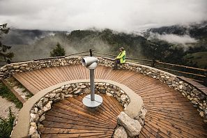 Wooden-clad panoramic platform in spiral form with stone wall and telescope on a mountain, a person leans on the railing and gazes into the misty mountain landscape below.