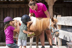 Zwei Kinder striegeln mit Unterstützung einer Betreuerin liebevoll ein Pony auf der Astberg Ponyalm in Going.