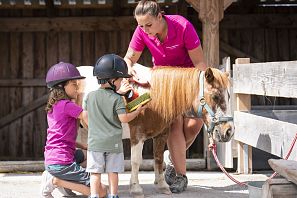 Zwei Kinder mit Reithelmen stehen neben einem kleinen braun-weißen Pony, begleitet von einer Betreuerin in pinkem Shirt, vor einem Holzstall.