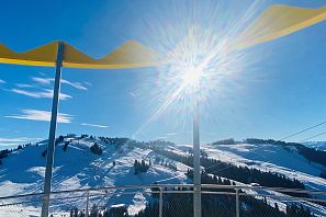 Blick von der Aussichtsplattform Kaiserliche Wohlfühlplattform in der SkiWelt Scheffau auf verschneite Hänge und Berge, Sonnenschein und das gelbe Kronendach im Vordergrund