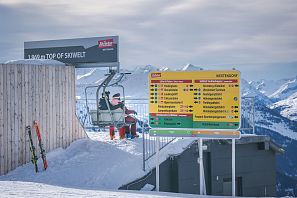 Skifahrer sitzen am Sessellift auf 1.869 Metern Höhe am höchsten Punkt der SkiWelt Westendorf; daneben bunter Wegweiser mit Pisten- und Liftübersicht vor verschneitem Alpenpanorama.