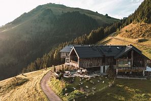 Moderne Berghütte mit Holzfassade in Hanglage, umgeben von grüner Almlandschaft und Wäldern, im warmen Licht der untergehenden Sonne