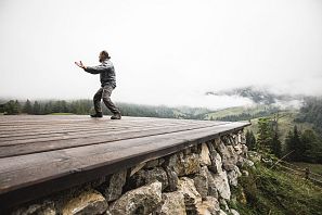 A person stands on a raised wooden platform with a stone foundation and performs mindful movements, with a misty mountain landscape with forests and meadows in the background.