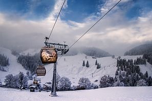 Gondeln einer Seilbahn fahren über eine verschneite Winterlandschaft mit bewaldeten Hängen und teils nebelverhangenem Himmel