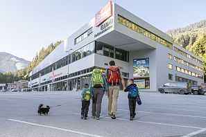 Family with two children and dog in the parking lot in front of the modern valley station building of the Hartkaiserbahn in Ellmau, surrounded by mountains in clear morning light.