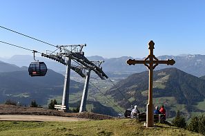 Gipfelkreuz mit Bank im Vordergrund, daneben drei Personen mit Blick ins Tal; daneben eine moderne 10er Gondelbahn Salvistabahn Itter mit Panoramaausblick auf grüne Hügel und Berge