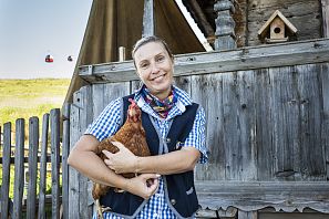 Lächelnde Frau hält Huhn in den Armen, steht vor einer alten Holzhütte mit Vogelhäuschen; im Hintergrund fahren Gondeln über eine Sommerwiese
