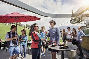 Gruppe fröhlicher Menschen genießt Getränke und Snacks auf einer Sonnenterrasse mit Blick auf grüne Hügel und eine Seilbahnstation