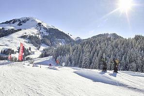 Verschneite Skipiste mit Funpark-Elementen und roten Fahnen im Skigebiet Söll, Kinder beim Skifahren, im Hintergrund verschneite Bäume und ein sonniger Himmel