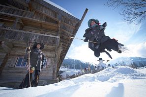 Zwei als Hexen verkleidete Personen im Schnee; eine springt mit Besen in der Luft, die andere steht lachend vor einem alten Holzhaus – winterliche Szenerie mit blauem Himmel