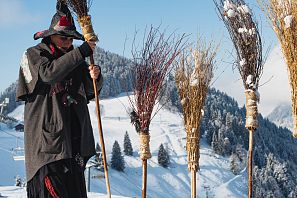 Frau in schwarz rotem Hexengewand steckt Holzbesen in den Schnee vor einem Schild: Frauen Parkplatz