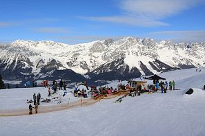 Winterliche Szene mit zahlreichen Skifahrern und Snowboardern rund um einen Snowpark-Bereich, im Hintergrund die schneebedeckten Gipfel des Kaisergebirges