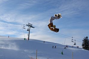 Ein Snowboarder führt einen spektakulären Rückwärtssalto vor blauem Himmel aus, im Hintergrund ein Skilift und mehrere Wintersportler auf der Piste