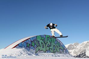 Ein Snowboarder balanciert auf einer farbig bemalten Rail mit Graffiti vor blauem Himmel und verschneiten Berggipfeln