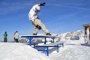 Ein Snowboarder führt einen Trick auf eine blau weißen Bank-Rail im Snowpark aus, mit schneebedeckten Bergen und blauem Himmel im Hintergrund