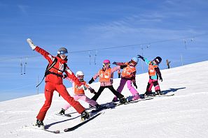 Ein Skilehrer und fünf Kinder in bunter Skikleidung fahren in einer Reihe eine sonnige Piste hinunter und breiten lachend die Arme aus