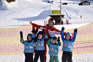 Skileherin steht im Skigebiet jubelnd hinter vier junge Skischüler welche über ihre Schikleidung hellblaue Warnwesten tragen