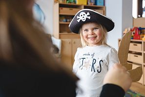 Lächelndes Mädchen mit Piratenhut und Kartonschwert steht in einem Raum mit Holzregalen und Spielzeug.
