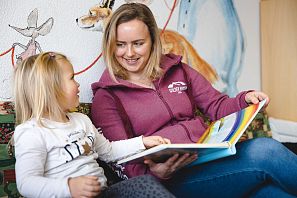 Erzieherin liest einem Mädchen auf einem Sofa ein Bilderbuch vor, bunte Tierzeichnungen schmücken die Wand im Hintergrund