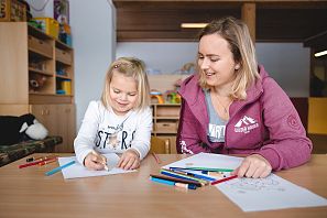 Ein Mädchen und eine Erzieherin sitzen an einem Tisch in der Kinderbetreuung und malen gemeinsam mit Buntstiften