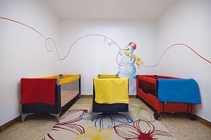 Drei farbige Kinderreisebetten stehen nebeneinander in einem hellen Raum mit Schneemann-Wandmalerei und bunter Blumenteppichdeko auf dem Boden.
