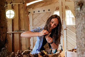 Frau bastelt konzentriert mit Naturmaterialien wie Holz und Zapfen in einer gemütlichen Holzhütte mit rundem Fenster und rustikalem Interieur.