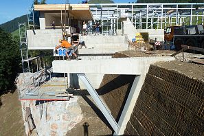 Mehrere Bauarbeiter arbeiten an einer im Rohbau befindlichen Bergbahnstation mit Stahl- und Betonkonstruktion, umgeben von grünem Bergpanorama