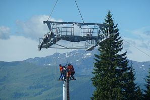 Mehrere Arbeiter in orangefarbener Sicherheitskleidung montieren ein großes Seilbahnbauteil mit einem Kran auf einer Stütze in alpiner Umgebung