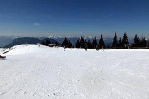 Extensa pista de esquí con nieve recién pisada bajo un cielo azul, estación de remonte con cabaña en primer plano y telesilla, así como laderas boscosas al fondo.