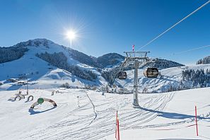 Verschneite Berglandschaft mit Gondelbahn, Skiparkour Hans im Glück und strahlendem Sonnenschein im Skigebiet, vor tiefblauem Himmel und bewaldeten Hängen