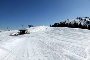 Extensa pista de esquí con nieve recién preparada bajo un cielo azul, estación de remonte con caseta en primer plano y telesilla, así como laderas boscosas al fondo.