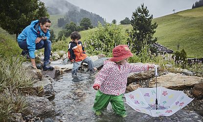 Zwei kleine Kinder spielen in wetterfester Kleidung in einem flachen Bachlauf, eines davon mit einem offenen Regenschirm im Wasser; ein Erwachsener beobachtet sie vom Rand aus