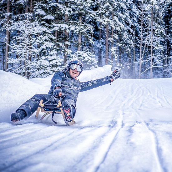 Zwei Personen rodeln lachend nebeneinander durch eine verschneite Winterlandschaft im Wald.
