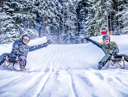 Zwei Personen rodeln lachend nebeneinander durch eine verschneite Winterlandschaft im Wald.