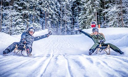 Zwei Personen rodeln lachend nebeneinander durch eine verschneite Winterlandschaft im Wald.