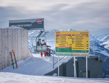 Skifahrer sitzen am Sessellift auf 1.869 Metern Höhe am höchsten Punkt der SkiWelt Westendorf; daneben bunter Wegweiser mit Pisten- und Liftübersicht vor verschneitem Alpenpanorama.