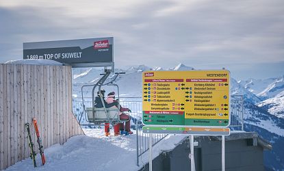 Skifahrer sitzen am Sessellift auf 1.869 Metern Höhe am höchsten Punkt der SkiWelt Westendorf; daneben bunter Wegweiser mit Pisten- und Liftübersicht vor verschneitem Alpenpanorama.