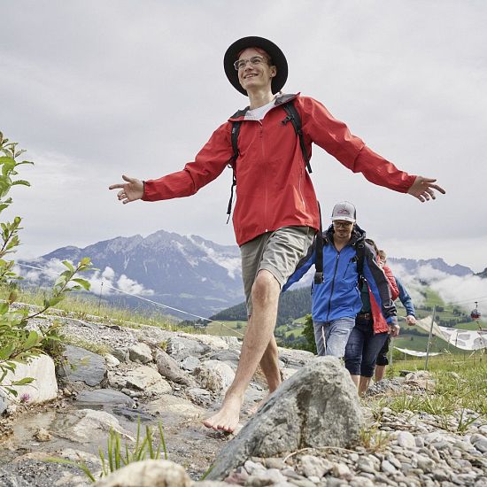 Barfußwanderer überqueren fröhlich einen Bach im alpinen Gelände mit Blick auf grüne Wiesen, Berge und Seilbahn in Tirol.