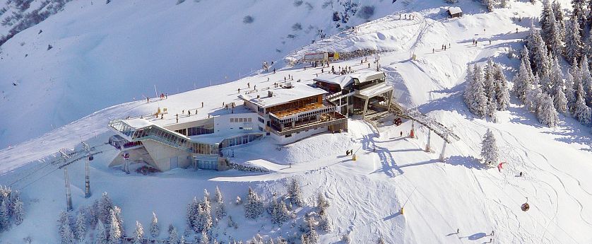 Verschneite Bergstation mit moderner Architektur im Skigebiet, umgeben von Skipisten, Gondeln und Skifahrern in winterlicher Hochgebirgslandschaft