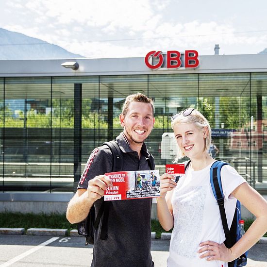 Junges Paar steht mit Hohe Salve Gästekarte vor einem ÖBB-Bahnhof in Tirol, im Hintergrund moderne Glasfassade und Berglandschaft.