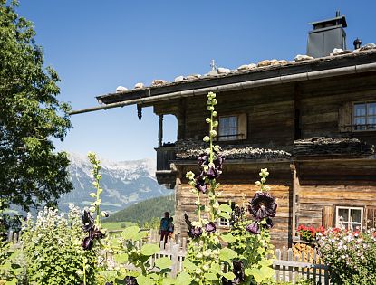 Holzhaus mit Schindeldach im traditionellen Stil, im Vordergrund blühende Stockrosen, dahinter Berglandschaft mit Wanderer im Hintergrund