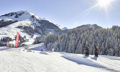 Verschneite Skipiste mit Funpark-Elementen und roten Fahnen im Skigebiet Söll, Kinder beim Skifahren, im Hintergrund verschneite Bäume und ein sonniger Himmel