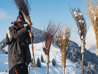 Frau in schwarz rotem Hexengewand steckt Holzbesen in den Schnee vor einem Schild: Frauen Parkplatz