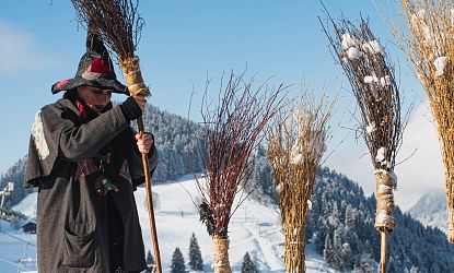 Frau in schwarz rotem Hexengewand steckt Holzbesen in den Schnee vor einem Schild: Frauen Parkplatz