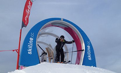 Skifahrer am Start einer Skiline-Rennstrecke, bereit zur Abfahrt durch ein rundes Starttor mit blau-weiß-roter Umrandung und Beschriftung „skiline.cc