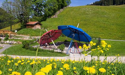 Zwei überdimensionale Sonnen­schirme – einer rot, einer blau – spenden Schatten auf einem modernen Spielplatz inmitten einer grünen Wiese mit blühenden Löwenzahnblumen und sanften Hügeln im Hintergrund