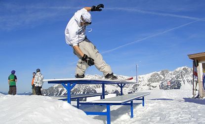 Ein Snowboarder führt einen Trick auf eine blau weißen Bank-Rail im Snowpark aus, mit schneebedeckten Bergen und blauem Himmel im Hintergrund