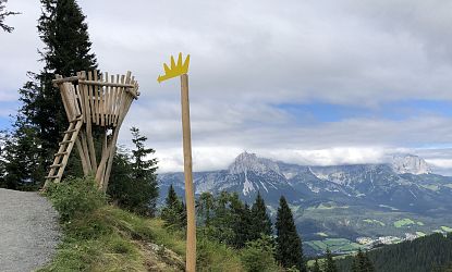 Kleine hölzerne Aussichtsplattform mit Leiter am Wegesrand, daneben ein Wegweiser mit gelber Krone. Im Hintergrund Panorama auf bewaldete Hügel und wolkenverhangene Gebirgskette